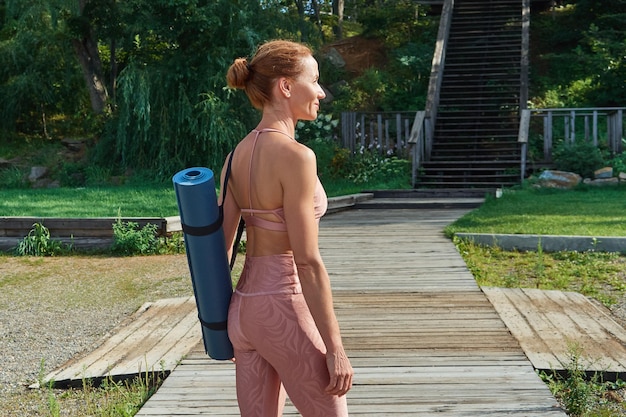 Profile view of a smiling woman in pink sportswear carries a mat standing on a wooden walkway about to do yoga practice outdoors