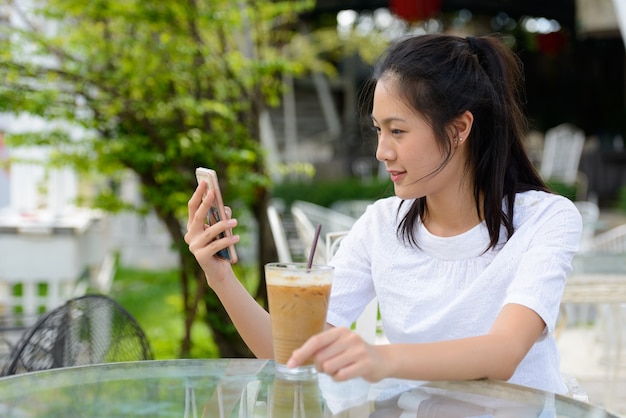 Profile view of young beautiful Asian woman using phone at the coffee shop outdoors