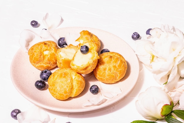 Profiteroles with blueberries. Custard cakes, Eclair, cream puff. Modern hard light, dark shadow. White peonies on plaster background, place for text