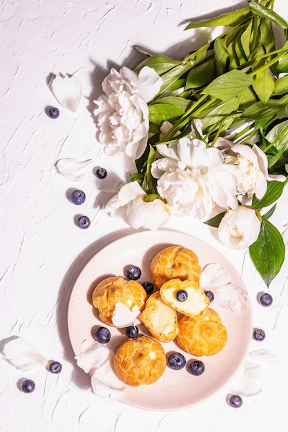 Profiteroles with blueberries. Custard cakes, Eclair, cream puff. Modern hard light, dark shadow. White peonies on plaster background, top view