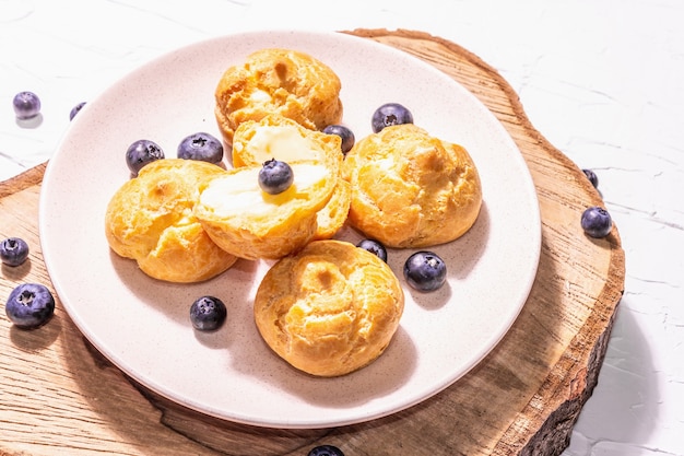 Profiteroles with blueberries. Custard cakes, Eclair, cream puff on a wooden stand, fashionable hard light, dark shadow. White plaster background, close up