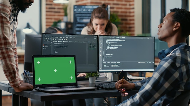 Programer taking glasses off talking with colleague at desk with laptop with green screen chroma key mockup and multiple computer screens compiling running code. Coders doing online cloud computing.