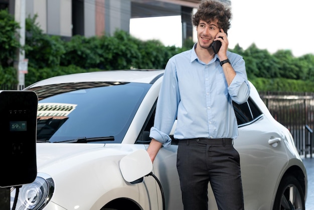 Progressive businessman talking on the phone with recharging electric vehicle