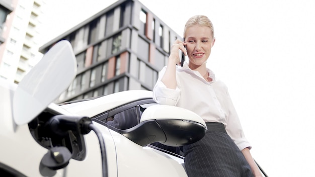 Progressive businesswoman leaning on electric car and charging station before driving around city center Eco friendly rechargeable EV car powered by sustainable and clean energy