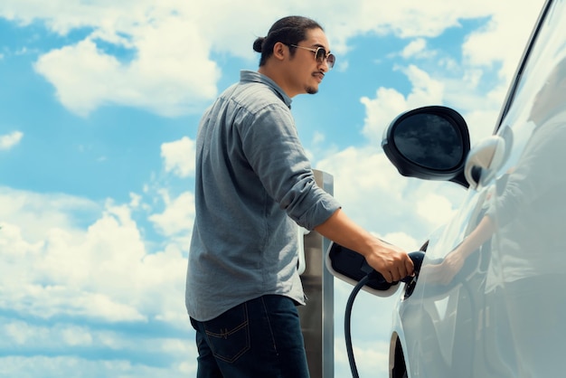 Progressive man with pluggedin EV car with charging point cloudscape backdrop