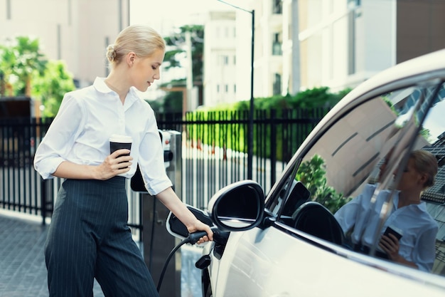 Progressive woman with coffee while charging EV car with residential buildings