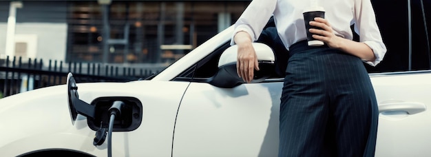 Progressive woman with coffee while charging EV car with residential buildings