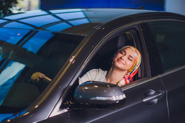 Property and people concept - muslim woman in hijab with car key over car show background. happy woman taking car key from dealer in auto show or salon