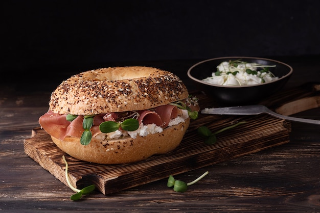 Prosciutto and cheese sandwich, cutting board with ham and ricotta bagels on a dark wooden background, rustic style.