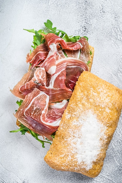 Prosciutto parma ham sandwich on ciabatta bread with arugula. White table. Top view.