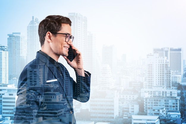 Prosperous handsome entrepreneur in suit and glasses pensively talking phone and looking on Bangkok cityscape The concept of problem solving consulting Downtown view Double exposure