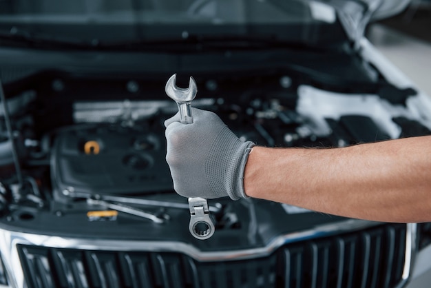 Protective gloves. Man's hand holds wrench in front of broken automobile.