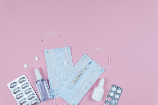 Protective medicine mask, antiseptic thermometer and pills on pink background, flat lay