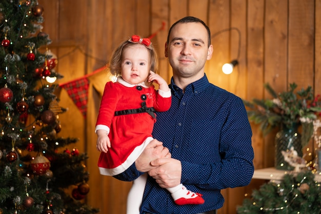 Proud dad holding his little daughter in his arms next to Christmas tree