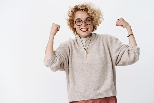 Proud and happy satisfied good-looking upbeat girl in glasses and sweater raising clenched fists in victory and celebration smiling from success and taste of win over white wall.