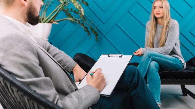 Psychologist writing notes on clipboard with pen during meeting with his female patient