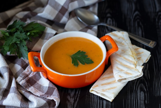 Pumpkin carrot cream soup in an orange plate with cheese pita on a checkered tablecloth with wooden background