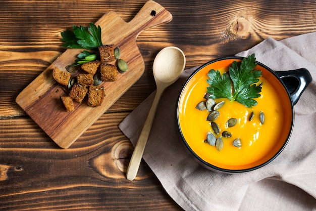 Pumpkin creamy soup in a bowl with seeds and parsley on a wooden background. Crackers with spices on the board. Copy space. Thanksgiving, autumn soup