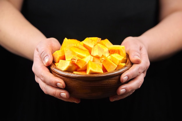 Pumpkin pieces in wooden bowl in woman hands on black background Chopped raw butternut squash