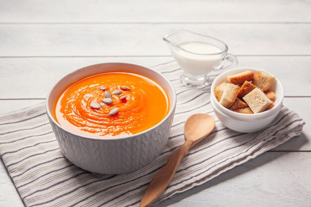 Pumpkin soup-puree with breadcrumbs, cream and seeds on towel, white wooden background.