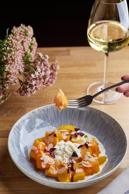 Pumpkin with stracciatella and microgreen on wooden table. Woman hold fork with pumpkin and glass of white wine on background