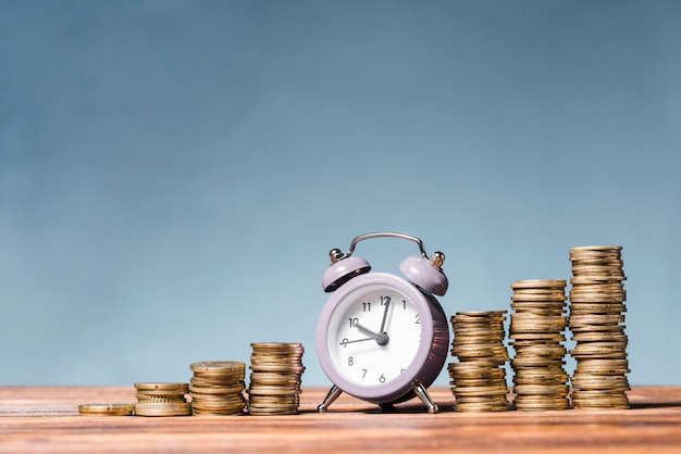 Purple alarm clock between the stack of increasing coins on wooden desk against blue background