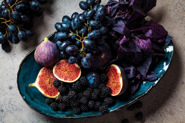 Purple fruits and berries in blue dish, gray background. Purple food background.