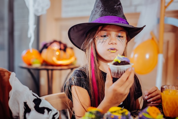 Purple lips. Funny cute dark-haired girl with purple and yellow lips wearing Halloween suit eating bright cupcake