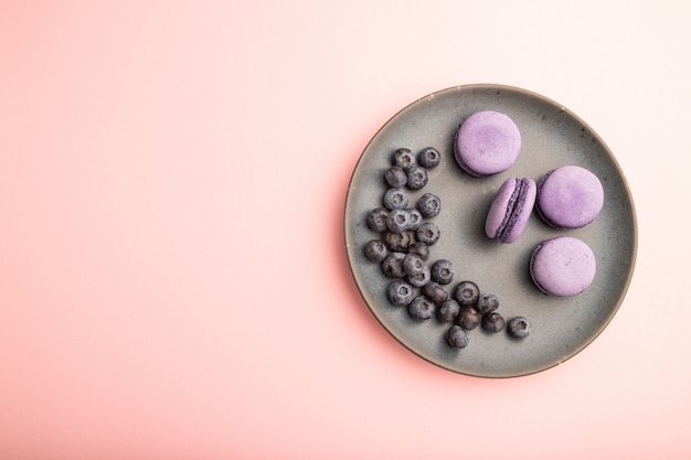 Purple macarons or macaroons cakes with blueberries on ceramic plate on a pastel pink background. 