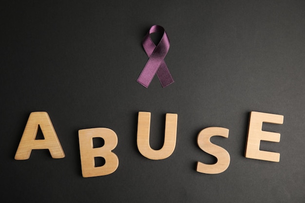 Purple ribbon and word ABUSE on black background flat lay Domestic violence awareness