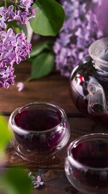 Purple tea is poured from a glass teapot into cups Lilac flowers are all around