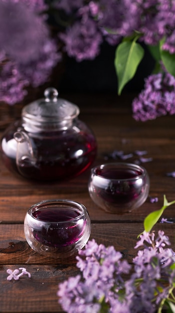 Purple tea is poured from a glass teapot into cups Lilac flowers are all around