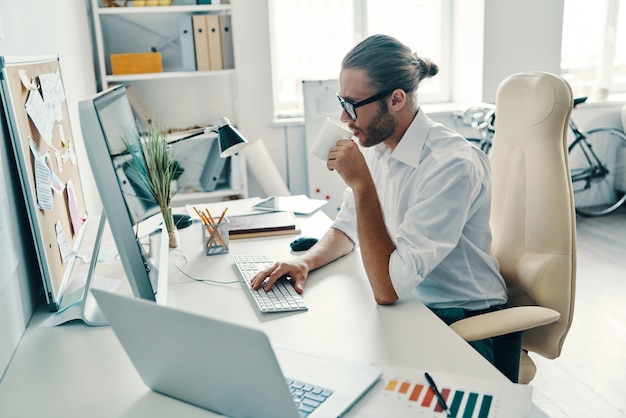 Putting ideas into something real. Thoughtful young man in shirt working using computer and drinking coffee while sitting in the office