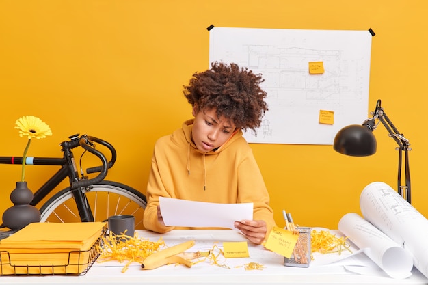 Puzzled busy Afro American female office worker has telephone conversation gets consultancy studies paper document works on design project poses at desktop with blueprints and sketches around