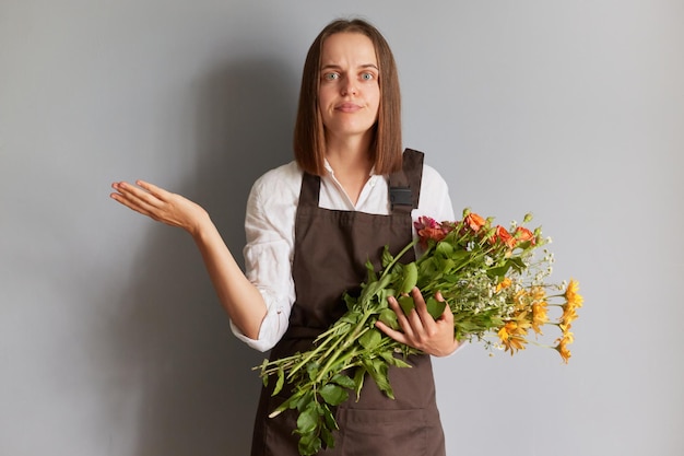 Puzzled confused female florist wearing brown apron holding flowers isolated over gray background spreading hand shrugging shoulders looking at camera with uncertain face