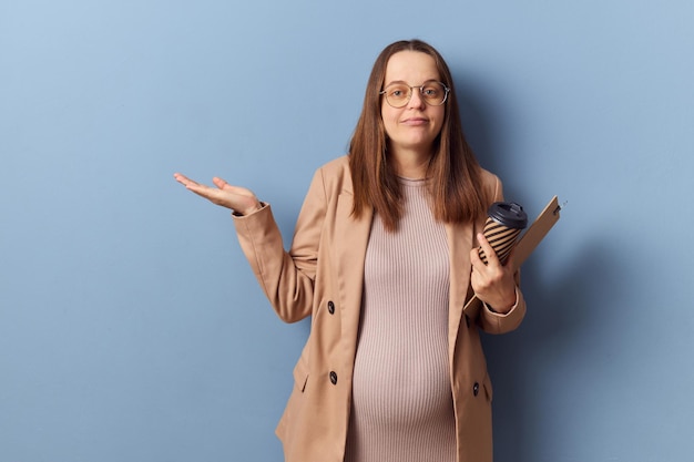 Photo puzzled confused pregnant woman wearing dress and jacket isolated over blue background holding documents and takeaway coffee shrugging shoulders with uncertain face