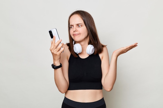 Puzzled confused slim woman with headphones wearing sportswear posing against gray background using mobile phone having problems with app