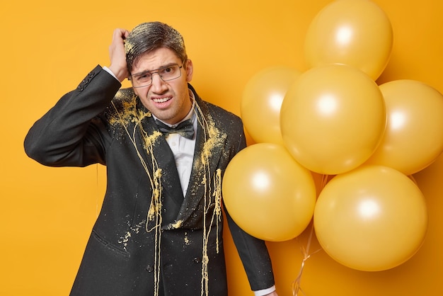 Puzzled displeased adult man scratches head looks frustrated wears transparent glasses and formal black suit holds inflated balloons isolated over yellow background Birthday celebration concept