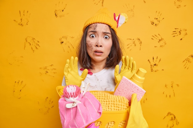 Puzzled excited Asian housewife stares at messy room doesnt know from what to begin raises hands in stop gesture wears hat rubber gloves poses near full laundry basket