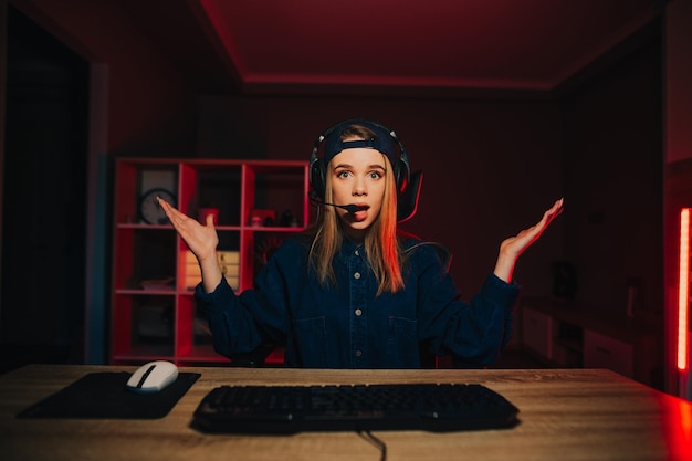 A puzzled female gamer in a headset sits at home in a room at night at a computer