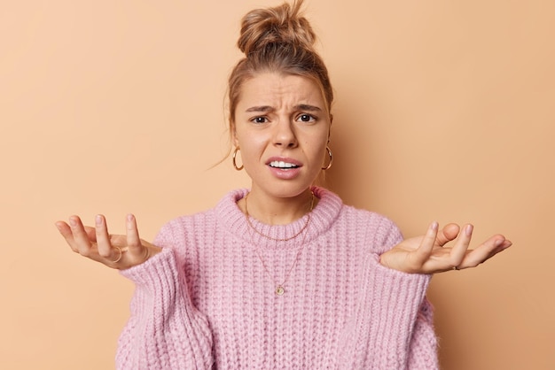 Puzzled indignant woman spreads palms feels hesitant looks displeased feels doubt cannot make decision wears knitted sweater isolated over beige background shrugs shoulders with bewilderment