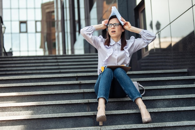 Puzzled tired business woman holds sheets of paper above her head while sitting on the stairs Lifestyle life concept