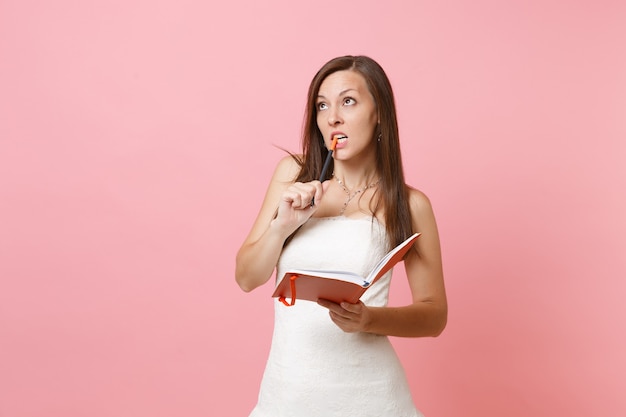 Puzzled woman in white dress gnawing pencil searching ideas, writing notes in diary, notebook