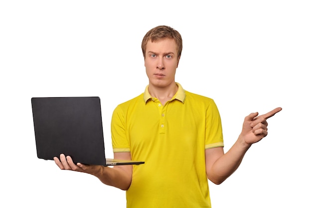 Puzzled young man in yellow Tshort holding laptop in black case and pointing with finger isolated on white background Funny confused man holding notebook in one hand Funny guy with bewildered face