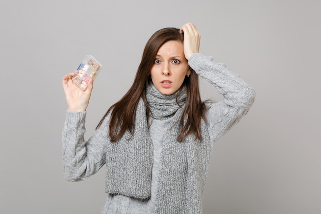 Puzzled young woman in gray sweater, scarf putting hand on head hold daily pill box isolated on grey background. Healthy lifestyle, ill sick disease treatment, cold season concept. Mock up copy space.