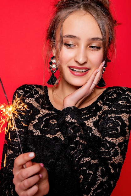 Pyrotechnics and people concept - smiling young woman or teenage girl happy woman with sparklers celebrate in black dress on red background. girl is surprised