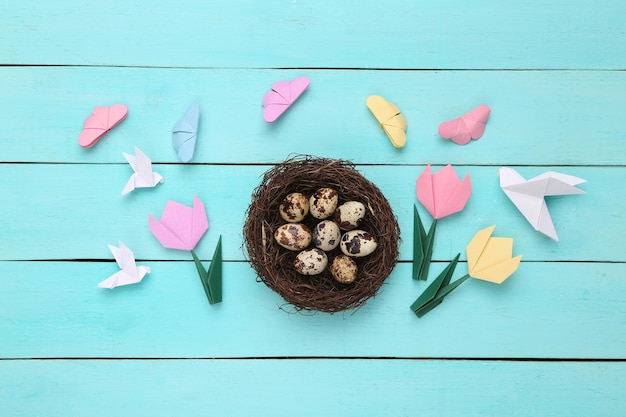 Quail eggs in a nest and origami butterflies tulips birds on blue wooden background Easter composition Top view