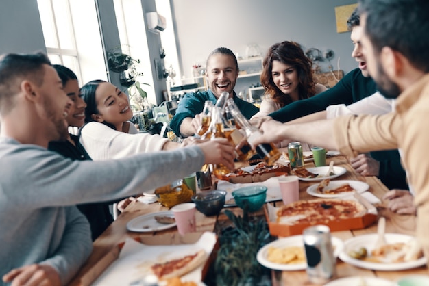 Quality time among friends. Group of young people in casual wear toasting each other and smiling while having a dinner party indoors