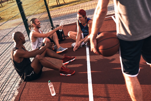 Quality time with friends. Top view of young men in sports clothing smiling while sitting on the basketball field outdoors