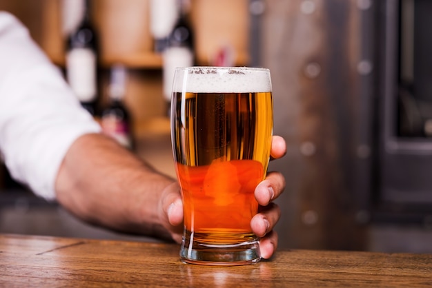 Quench your thirst with glass of cold beer! Close-up of man stretching out glass with beer while standing at the bar counter